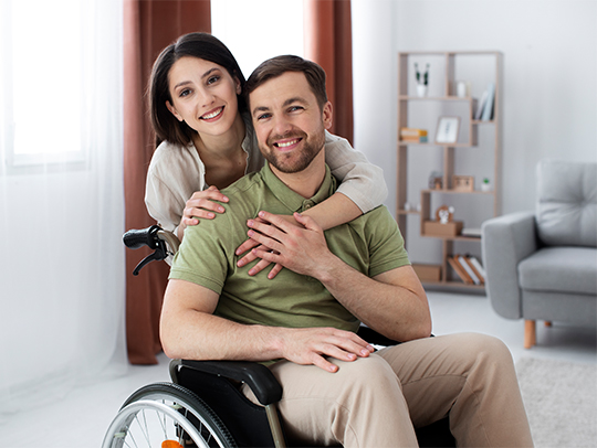 Smiling man in a wheelchair embraced by a supportive woman in a cozy home, highlighting NDIS disability services Melbourne.