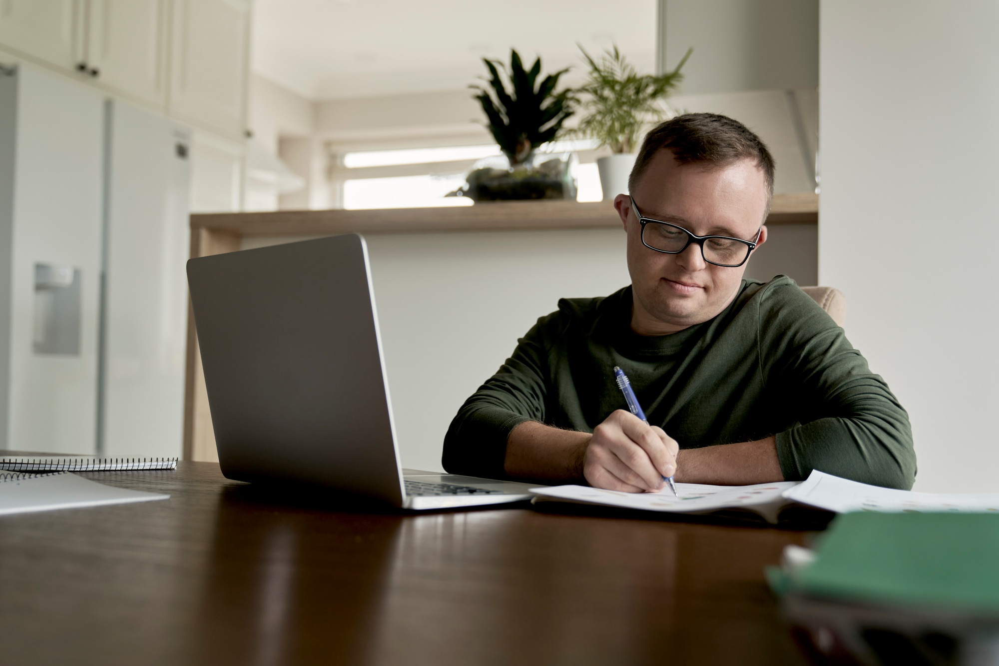 Person working on a laptop and writing in a notebook at home, representing NDIS education support for learning and employment goals.